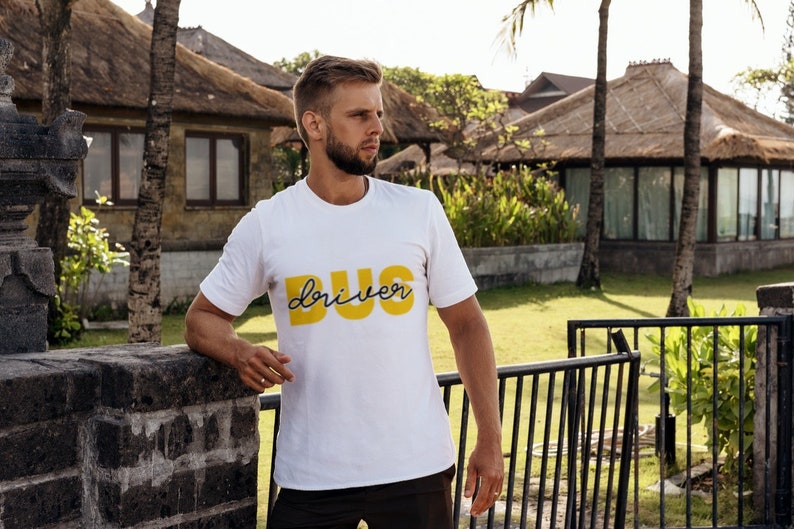 Puede incluir: Un hombre con una camiseta blanca con las palabras "BUG driver" impresas en amarillo en la parte delantera. Est&aacute; de pie frente a una puerta y mirando hacia la derecha.