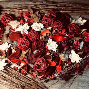 Op de afbeelding: Een hartvormige opstelling van potpourri met rode en witte gedroogde bloemen, rode kaneelstokjes en bruine takjes. De potpourri is op een houten oppervlak gerangschikt.
