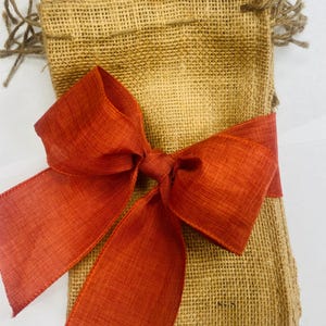May include: A stack of rectangular tan burlap pouches featuring a coarse woven texture and frayed edges at the top. The pouches are tied together with a wide, vibrant burnt orange ribbon that is knotted into a large, prominent bow at the centre.