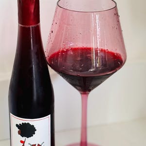 May include: A bottle of red wine with a label featuring a cartoon illustration of a person with an afro. The bottle is next to a glass of red wine in a pink glass.