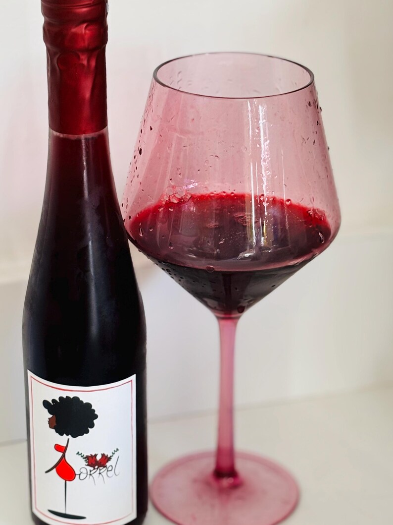 May include: A bottle of red wine with a label featuring a cartoon illustration of a person with an afro. The bottle is next to a glass of red wine in a pink glass.