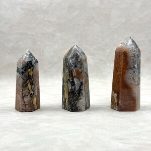 Puede incluir: Tres obeliscos de piedra pulida con diversos patrones y colores. Las piedras presentan tonos marrones, grises, negros y crema, con vetas visibles y texturas naturales. Cada obelisco tiene una parte superior puntiaguda y una base rectangular.