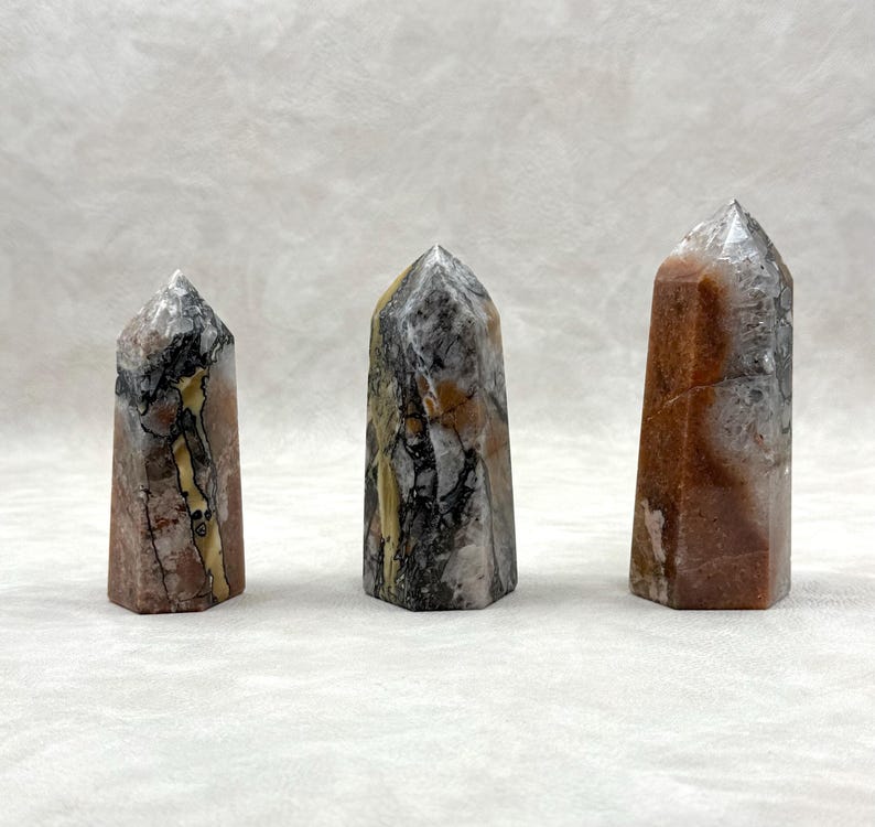 Puede incluir: Tres obeliscos de piedra pulida con diversos patrones y colores. Las piedras presentan tonos marrones, grises, negros y crema, con vetas visibles y texturas naturales. Cada obelisco tiene una parte superior puntiaguda y una base rectangular.