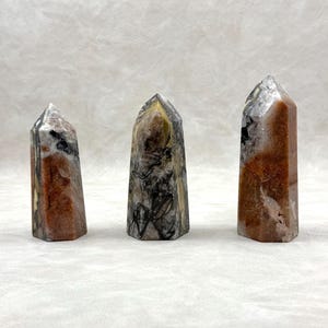 Puede incluir: Tres torres de piedras preciosas pulidas con una mezcla de colores que incluyen rojo, marr&oacute;n, negro y blanco. Los cristales tienen una parte superior puntiaguda y una base rectangular. Los cristales se exhiben sobre una superficie blanca.