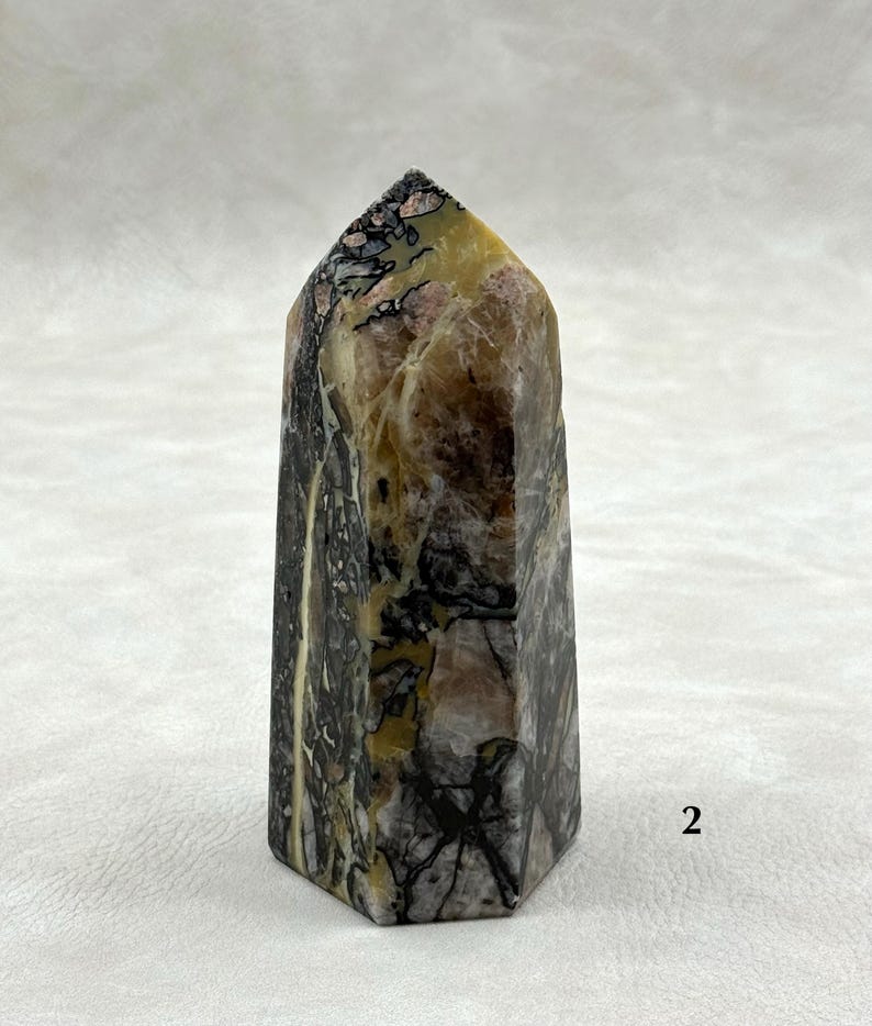 Puede incluir: Un obelisco de piedra hexagonal con una parte superior puntiaguda. La piedra presenta una mezcla de colores, incluyendo negro, marr&oacute;n, amarillo y rosa, con intrincados patrones y vetas. El obelisco es un art&iacute;culo decorativo.