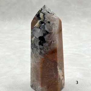 Puede incluir: Una punta de cristal hexagonal con una superficie pulida. El cristal presenta una mezcla de colores, incluyendo marr&oacute;n, negro, blanco y cuarzo transparente. La parte superior es puntiaguda. El cristal es una piedra natural.