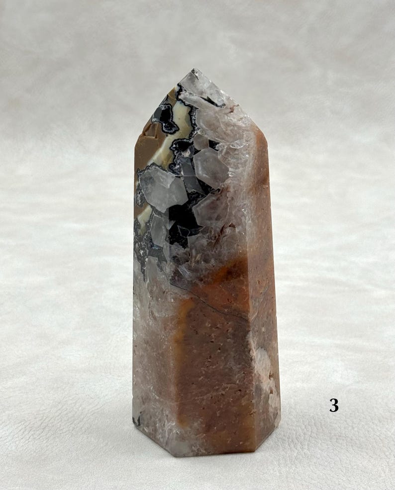 Puede incluir: Una punta de cristal hexagonal con una superficie pulida. El cristal presenta una mezcla de colores, incluyendo marr&oacute;n, negro, blanco y cuarzo transparente. La parte superior es puntiaguda. El cristal es una piedra natural.