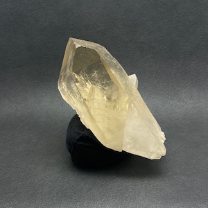 Puede incluir: Un gran cristal de cuarzo translúcido con un tono amarillo claro. El cristal está montado sobre una pequeña base de terciopelo negro, sobre un fondo oscuro. Las facetas del cristal capturan la luz, mostrando su forma natural.