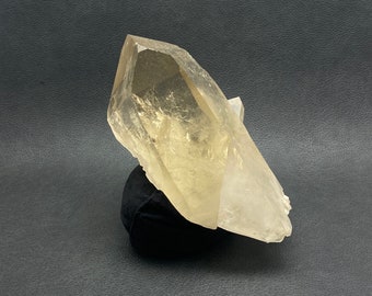 May include: A large, translucent quartz crystal with a light yellow hue. The crystal is mounted on a small, black velvet base, set against a dark background. The crystal's facets catch the light, showcasing its natural form.