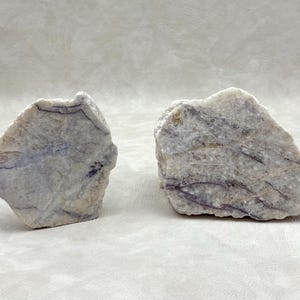 May include: Two raw, uncut stones with a marbled appearance. The stones are primarily white with streaks of light purple and gray. The stones have a rough, natural texture and irregular shapes, suggesting they are natural mineral specimens.