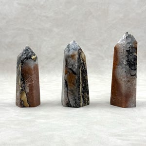 Puede incluir: Tres obeliscos de piedra pulida en tonos marr&oacute;n, gris, negro y crema. Cada obelisco tiene un patr&oacute;n y una forma &uacute;nicos, con una parte superior puntiaguda. Las piedras se exhiben sobre un fondo neutro.