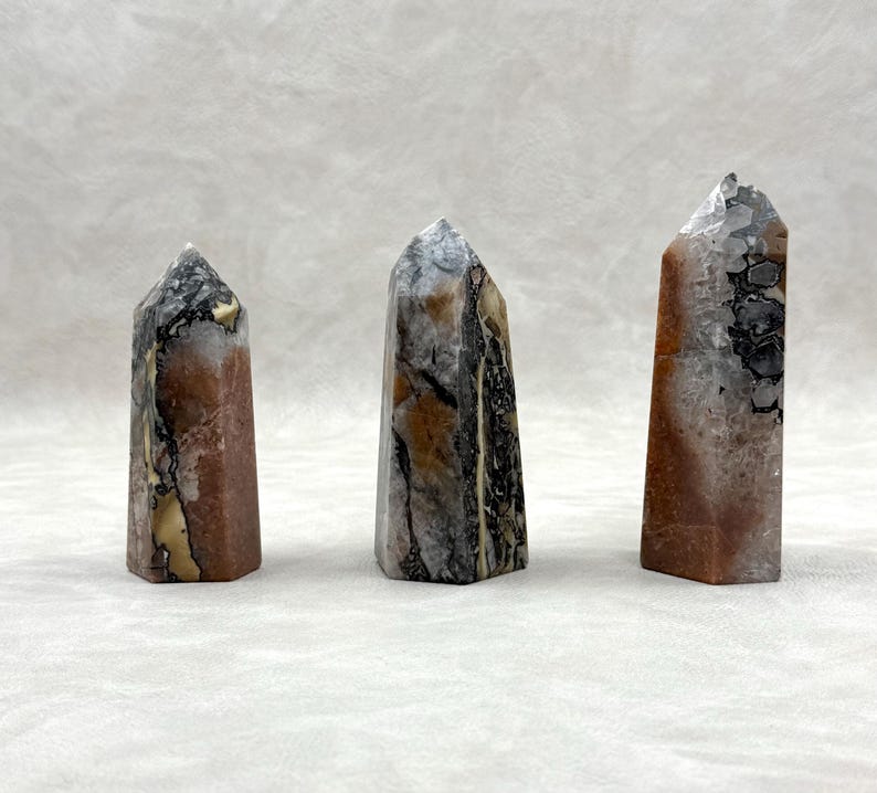 Puede incluir: Tres obeliscos de piedra pulida en tonos marr&oacute;n, gris, negro y crema. Cada obelisco tiene un patr&oacute;n y una forma &uacute;nicos, con una parte superior puntiaguda. Las piedras se exhiben sobre un fondo neutro.