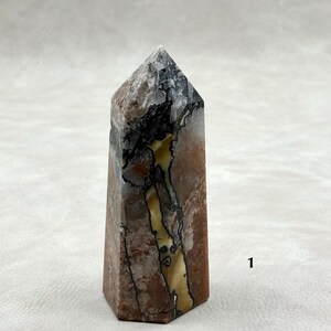 Puede incluir: Una punta de cristal hexagonal con una parte superior plana. La piedra presenta una mezcla de colores, incluyendo marr&oacute;n, negro, blanco y amarillo. El cristal tiene una superficie pulida y se muestra sobre un fondo neutro. Aproximadamente 10 cm de alto.