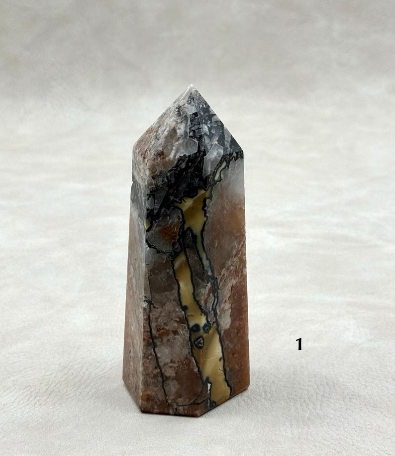 Puede incluir: Una punta de cristal hexagonal con una parte superior plana. La piedra presenta una mezcla de colores, incluyendo marr&oacute;n, negro, blanco y amarillo. El cristal tiene una superficie pulida y se muestra sobre un fondo neutro. Aproximadamente 10 cm de alto.