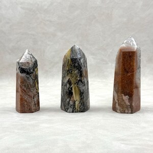 Puede incluir: Tres obeliscos de piedras preciosas pulidas. Los obeliscos son una mezcla de colores, incluyendo marr&oacute;n, gris y blanco, con una apariencia cristalina. Cada obelisco tiene una parte superior puntiaguda y una base rectangular. Las piedras preciosas se exhiben sobre una superficie blanca.