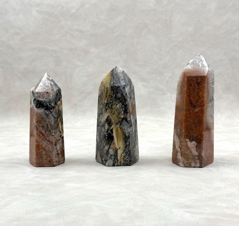 Puede incluir: Tres obeliscos de piedras preciosas pulidas. Los obeliscos son una mezcla de colores, incluyendo marr&oacute;n, gris y blanco, con una apariencia cristalina. Cada obelisco tiene una parte superior puntiaguda y una base rectangular. Las piedras preciosas se exhiben sobre una superficie blanca.