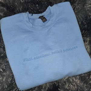May include: A light blue sweatshirt with the embroidered text "What excellent boiled potatoes". The crew neck sweatshirt is made of a soft material and is a solid light blue color.