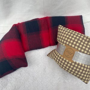 May include: A red and black plaid neck pillow and a brown and beige gingham pillow with a "Stitched By Dawn" label. Both pillows are on a white surface.