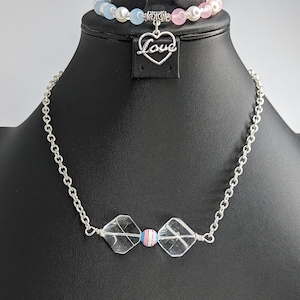 May include: A silver chain necklace with two clear crystal beads and a single blue, pink, and white bead in the center. The necklace is on a black mannequin with a silver heart charm that says "Love".