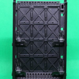 May include: A black plastic structure with a geometric design. The structure has a rectangular shape with a grid-like pattern of intersecting lines and square details. It features vertical supports and a base, likely for display or support.