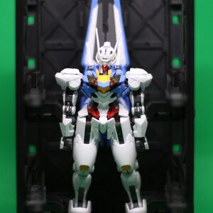 May include: A detailed action figure of a robot, with a white, blue, red, and yellow colour scheme. The robot stands in a black display case against a green backdrop. The figure has articulated joints and intricate detailing.