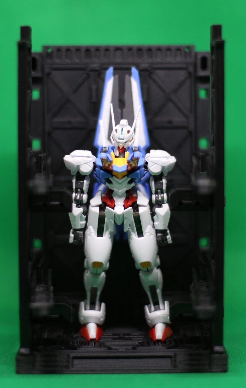 May include: A detailed action figure of a robot, with a white, blue, red, and yellow colour scheme. The robot stands in a black display case against a green backdrop. The figure has articulated joints and intricate detailing.