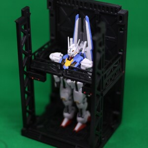 May include: A detailed robot model in white, blue, and yellow is positioned within a black geometric structure. The robot features red feet and white armour. The structure has a cage-like design with support beams and a platform.