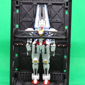 May include: A detailed robot action figure in white, blue, red, and yellow is presented within a black, rectangular, 3D-printed display case. The robot has articulated joints and is posed standing. The background is a solid green.