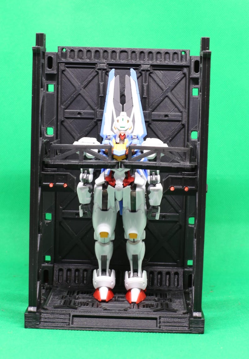 May include: A detailed robot action figure in white, blue, red, and yellow is presented within a black, rectangular, 3D-printed display case. The robot has articulated joints and is posed standing. The background is a solid green.