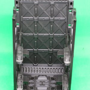 May include: A black, rectangular plastic object with a detailed geometric design. The object has a series of rectangular openings at the bottom and a grid-like pattern on the main surface. It features several protruding elements and is set against a green background.