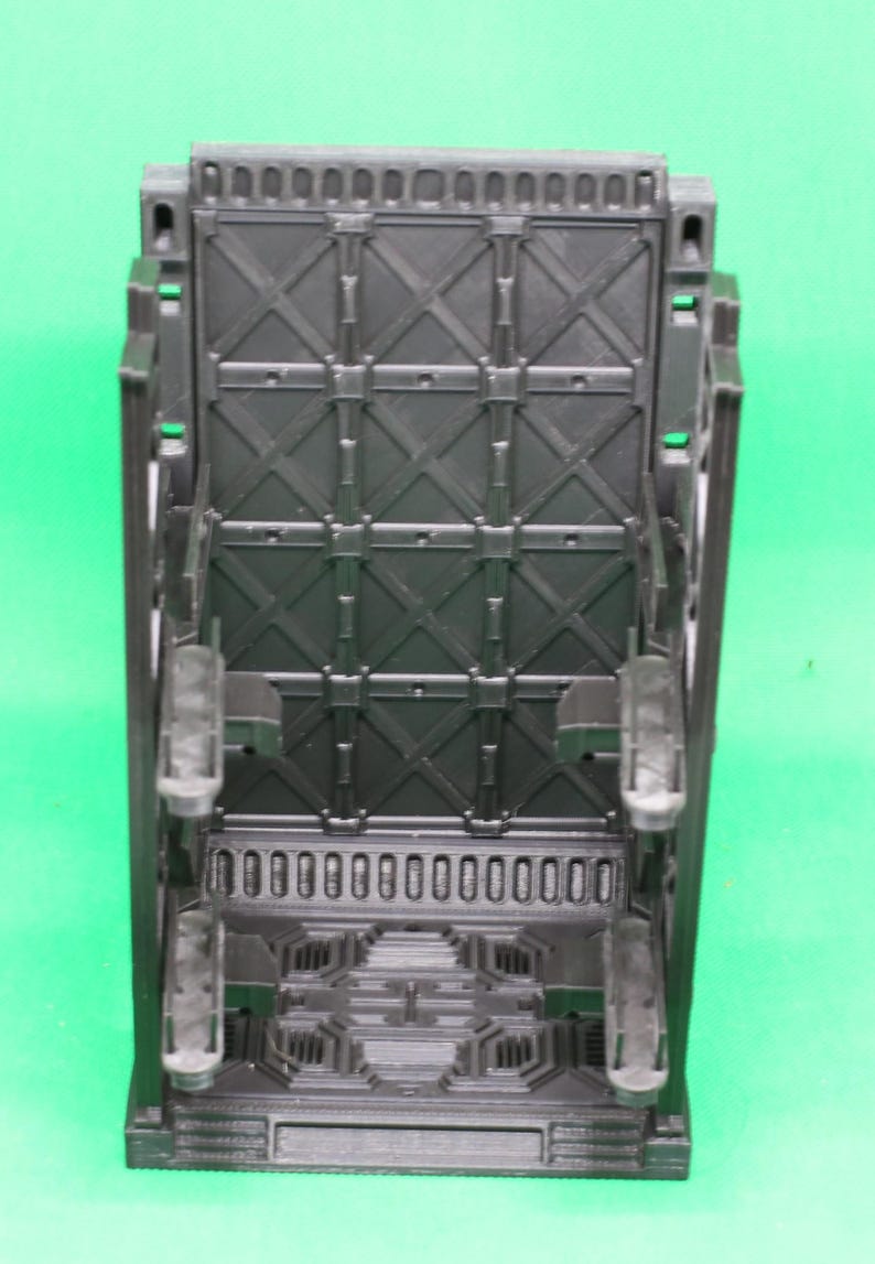 May include: A black, rectangular plastic object with a detailed geometric design. The object has a series of rectangular openings at the bottom and a grid-like pattern on the main surface. It features several protruding elements and is set against a green background.