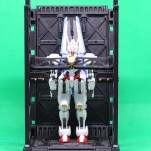 May include: A white, blue, red, and yellow action figure stands inside a black, rectangular display case. The figure has a detailed design with articulated joints. The display case has a series of arched cutouts along the top edge.