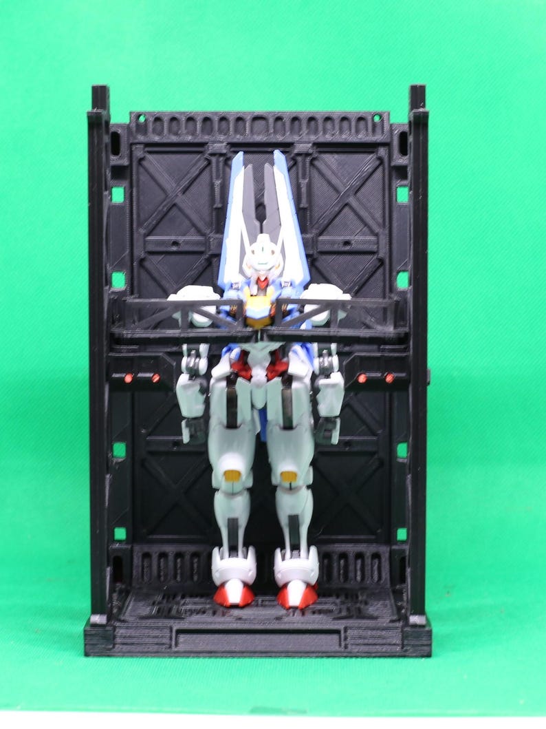 May include: A white, blue, red, and yellow action figure stands inside a black, rectangular display case. The figure has a detailed design with articulated joints. The display case has a series of arched cutouts along the top edge.