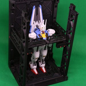 May include: A detailed robot figure in white, blue, and red, positioned within a black geometric structure. The robot features a yellow chest and red feet. The structure has an industrial design with multiple levels and support beams.