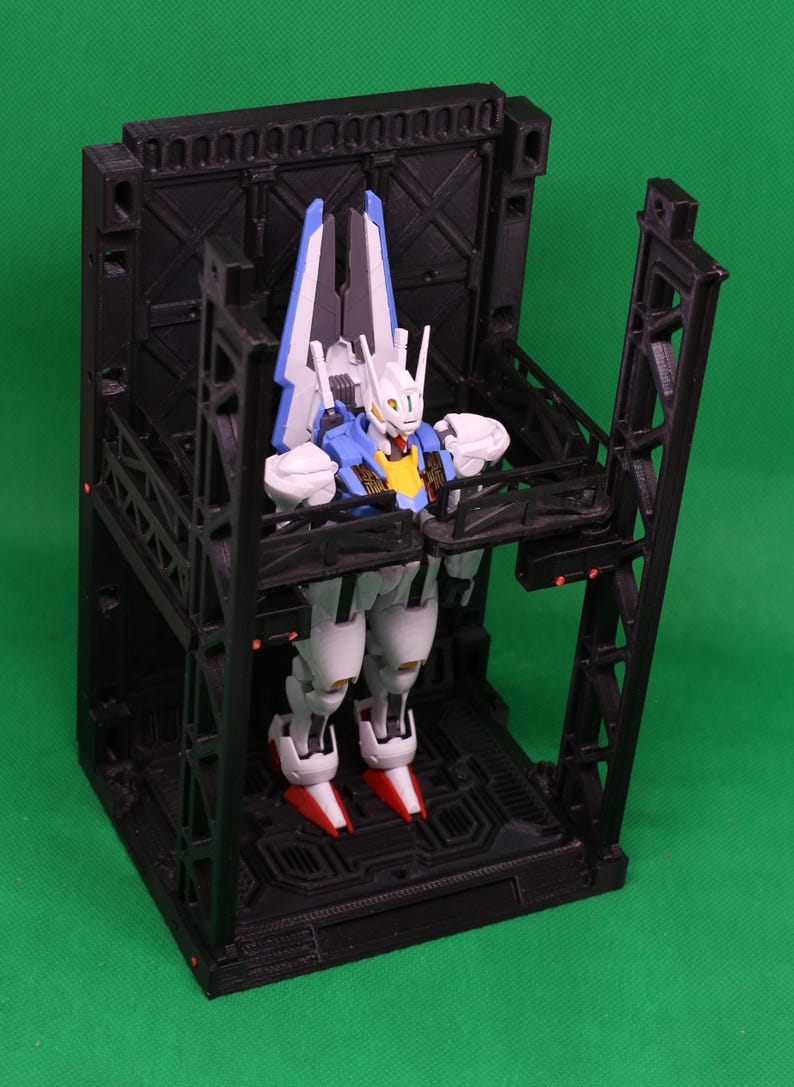 May include: A detailed robot figure in white, blue, and red, positioned within a black geometric structure. The robot features a yellow chest and red feet. The structure has an industrial design with multiple levels and support beams.
