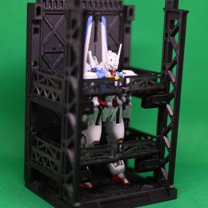 May include: A white, blue, and red action figure is positioned within a black, multi-level display case. The figure has a detailed design with a helmet and wings. The display case has a geometric design and is set against a green backdrop.