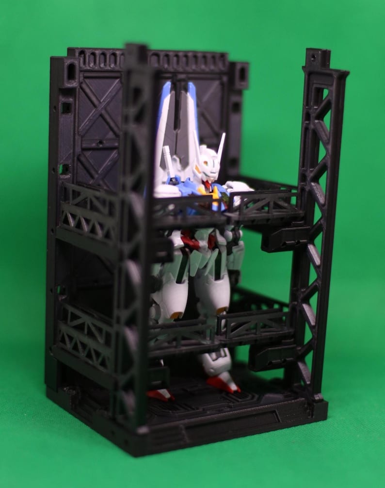 May include: A white, blue, and red action figure is positioned within a black, multi-level display case. The figure has a detailed design with a helmet and wings. The display case has a geometric design and is set against a green backdrop.