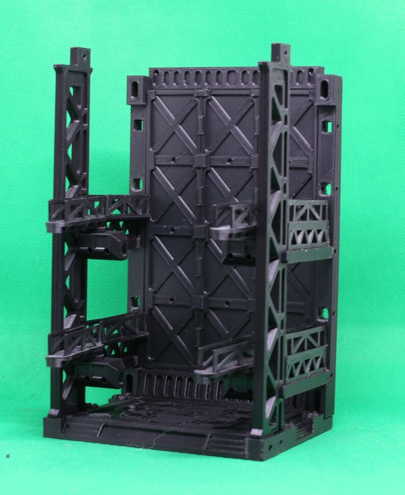 May include: A black, modular diorama set with an industrial design. The set features multiple levels, walkways, and a central wall with an X-shaped pattern. The structure is made of plastic and is designed for miniature figures.