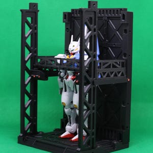 May include: A black plastic model kit of a mech robot figure inside a black structure. The robot is white, red, blue, and yellow. The structure has multiple levels and support beams, set against a green backdrop.