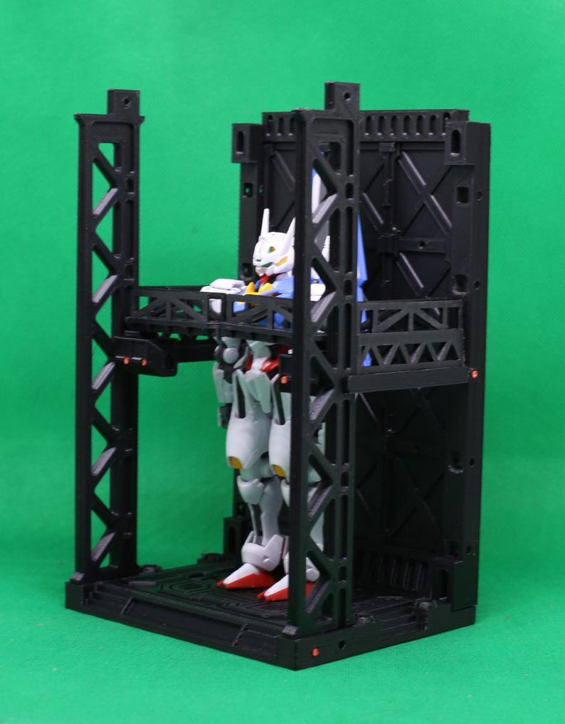 May include: A black plastic model kit of a mech robot figure inside a black structure. The robot is white, red, blue, and yellow. The structure has multiple levels and support beams, set against a green backdrop.