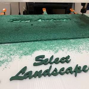 May include: Close-up of a CNC machine cutting out the words "Select Landscape" from a green sheet of material. The cut-out letters are on a white surface with green shavings.