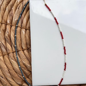 May include: A beaded necklace with a gold clasp. The necklace features three distinct sections: a dark blue section with small white beads, and a section with alternating red and white beads. The necklace is displayed on a woven surface.