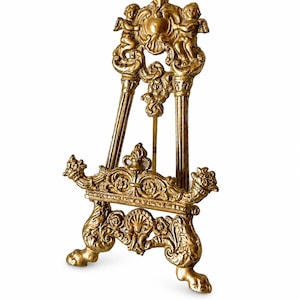 May include: A gold-colored metal easel stand featuring ornate relief carvings of cherubs, scrollwork, and shell motifs. The structure includes two vertical supports, a horizontal ledge for display, and a tripod base with decorative feet.
