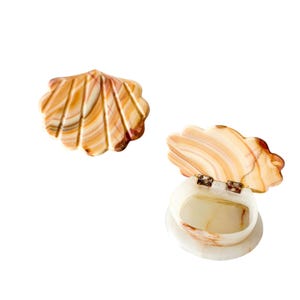 May include: Two seashell-shaped trinket boxes. One is closed, the other is open, revealing the interior. The boxes are made of a cream-colored material with brown and orange veining. The open box has small metal hinges.