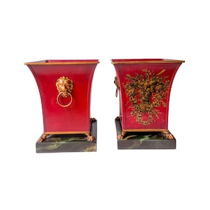 May include: Pair of red decorative planters with gold lion head handles and feet. One planter features a floral design. The planters sit on a dark green and black base. The planters are a square shape.