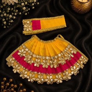 May include: A yellow and hot pink pleated skirt and a matching rectangular headband, both featuring gold metallic trim and decorative flower-shaped mirror embellishments along the edges. The set is designed with vibrant fabric layers and reflective accents.