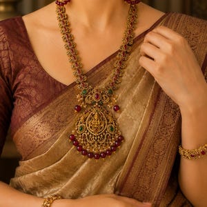 May include: A long gold-toned necklace featuring intricate metalwork, embedded with small red and green stones, and accented by dangling deep red beads. The central pendant displays a detailed embossed figure, complemented by matching gold-toned bracelets.
