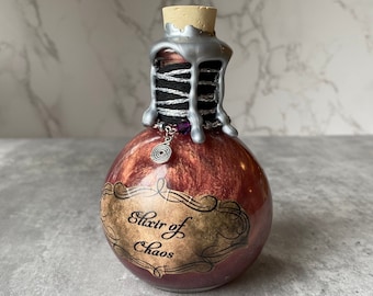 May include: A round, red-brown glass bottle with a cork stopper and silver wax seal. The bottle has a label that reads "Elixir of Chaos" in a vintage font. A silver charm and black twine detail the neck.