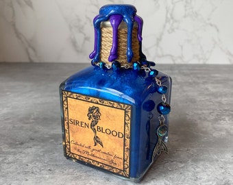 May include: A blue glass bottle with a purple and blue wax seal. The bottle has a label that reads "SIREN BLOOD Collected with great caution from the Mediterranean Sea".
