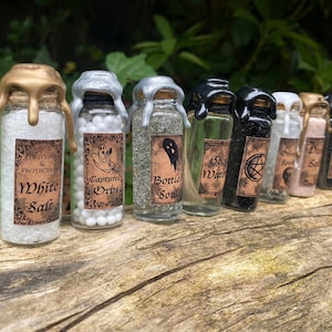 May include: Eight small glass bottles with cork stoppers and labels. The bottles are filled with various substances, including white salt, white beads, glitter, clear liquid, black sand, and pink salt. The labels have text such as "Purifying & Protection", "White Salt", "Captured Orbs", "Bottled Souls", "Gold Water", "Spirit", and "Pink Salt".
